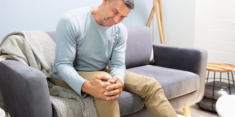 An elderly man sitting in a chair, looking distressed while holding his knee in pain.